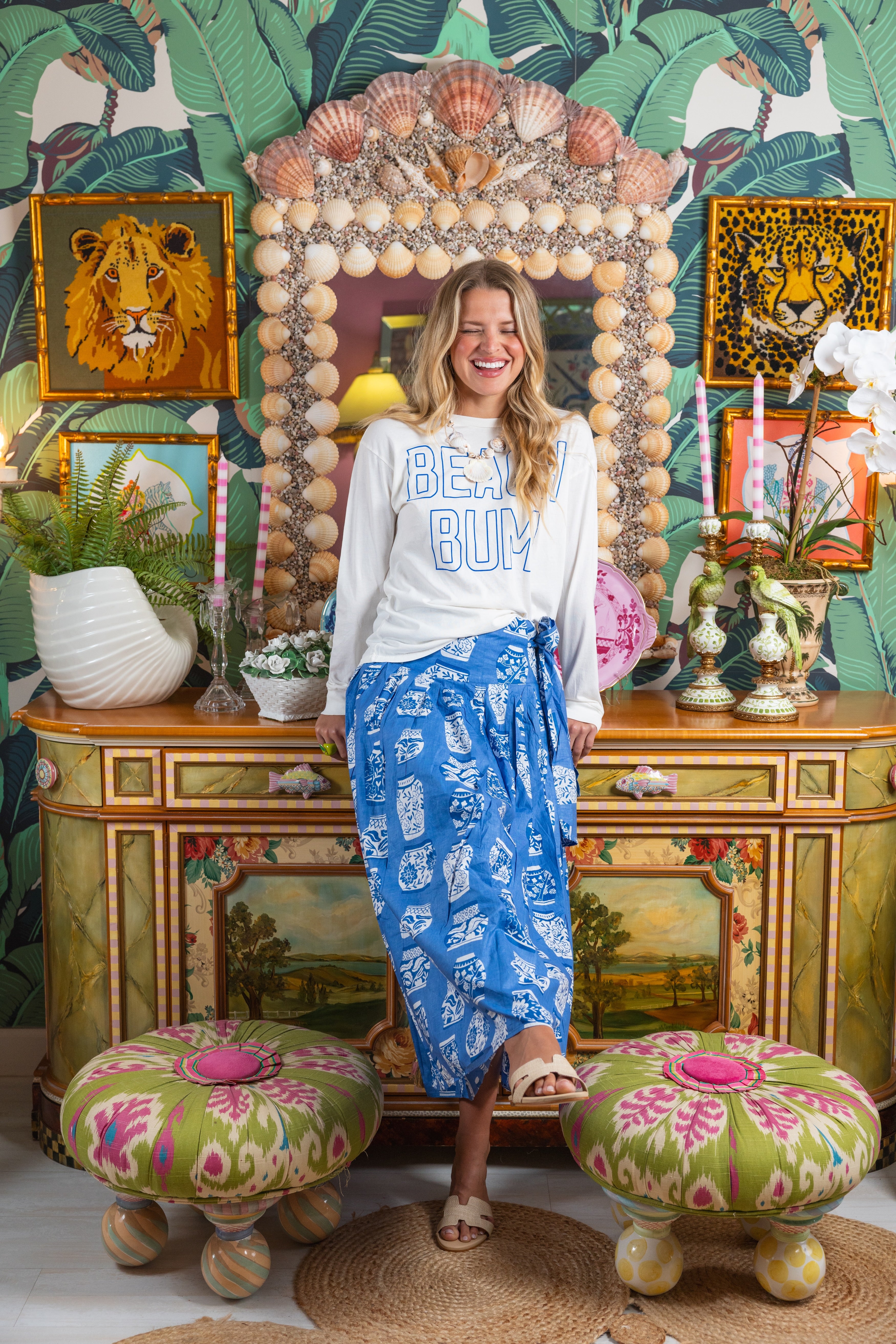 Woman standing in a decorated room with a shell-framed mirror and colorful decor.
