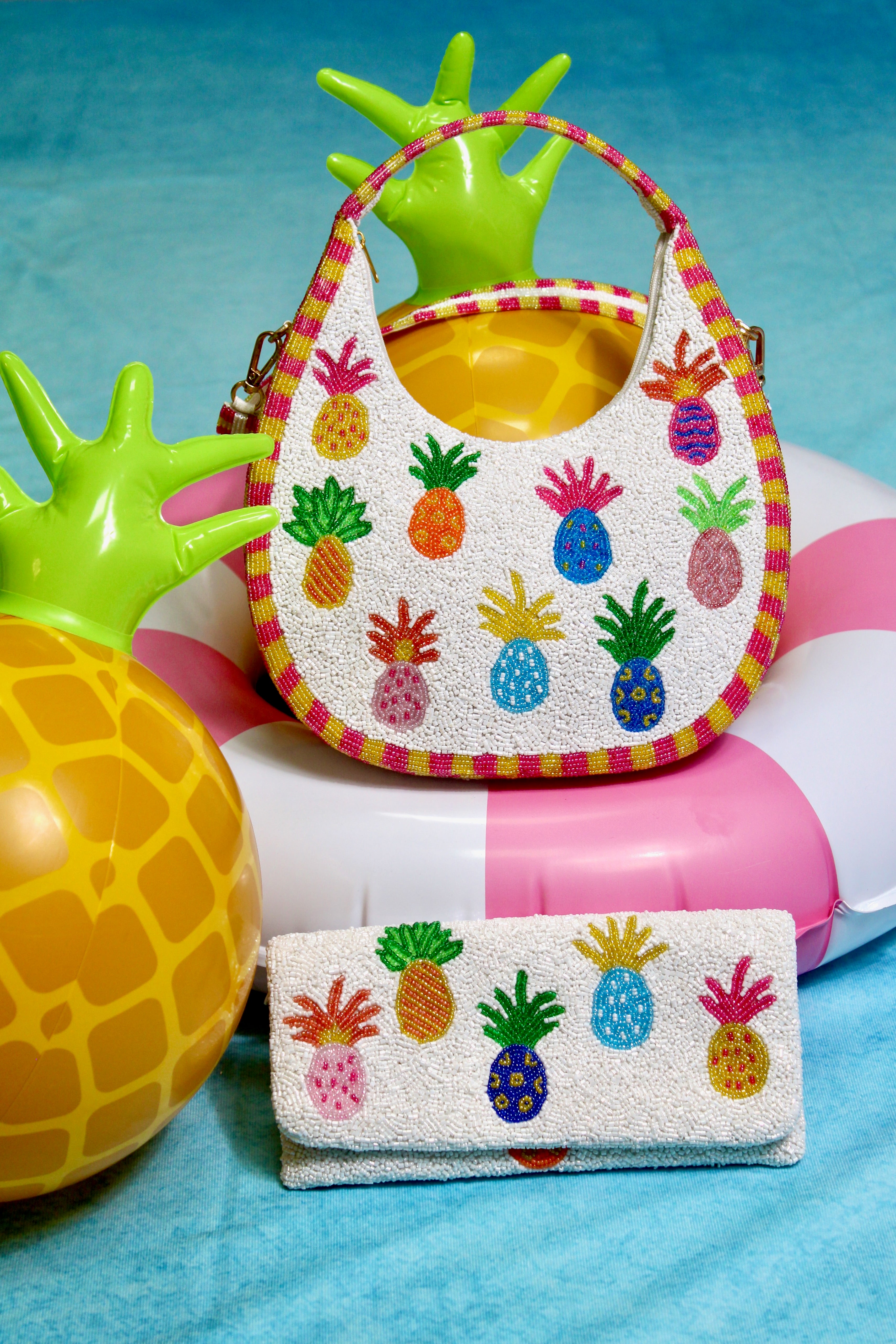Colorful pineapple-patterned handbag and wallet on a beach setting with inflatable pineapple and palm tree.