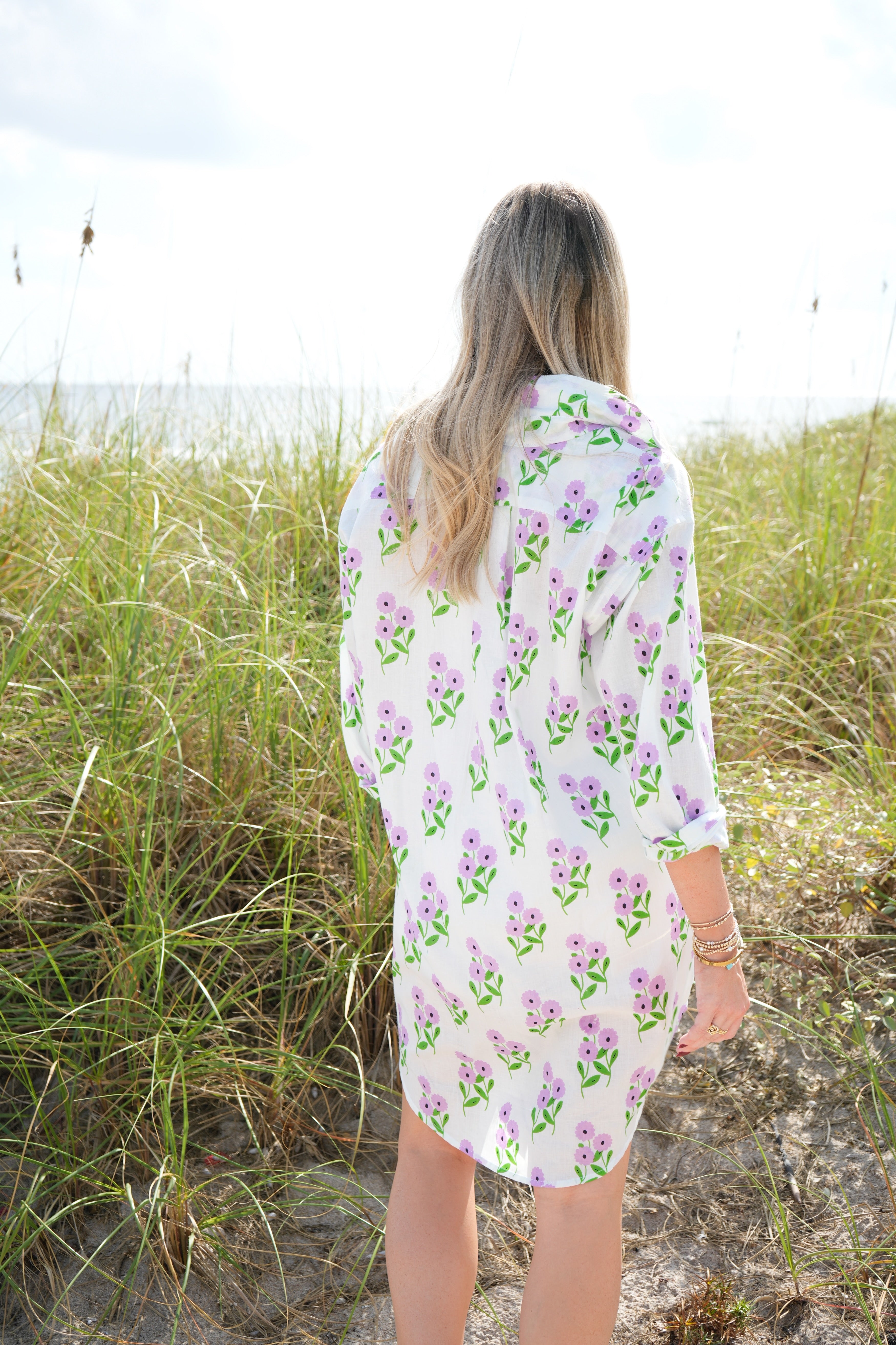 Woman wearing a floral dress standing on a beach with grass and sand.