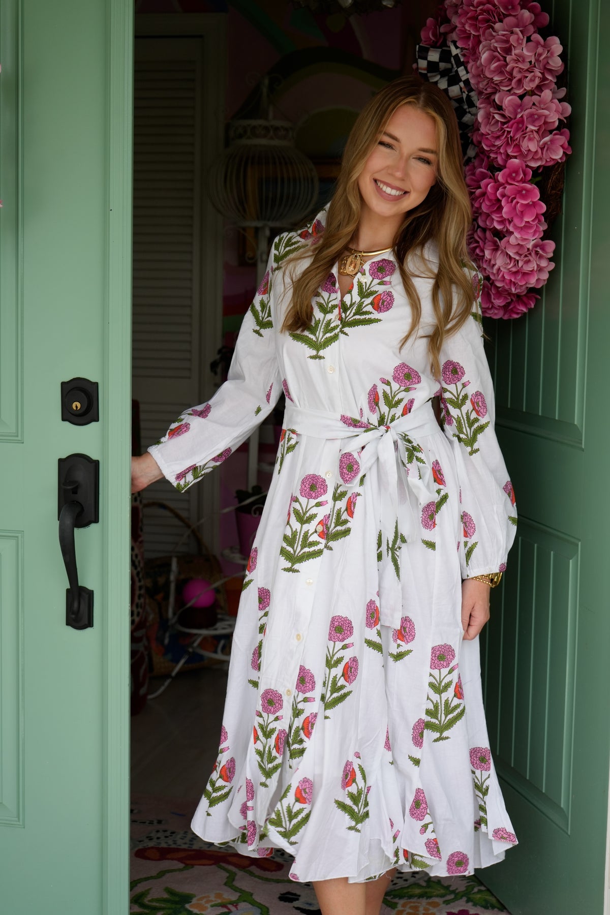 A woman wearing a white long-sleeve midi dress with a pink and green floral print, standing in a doorway of a mint green door.
