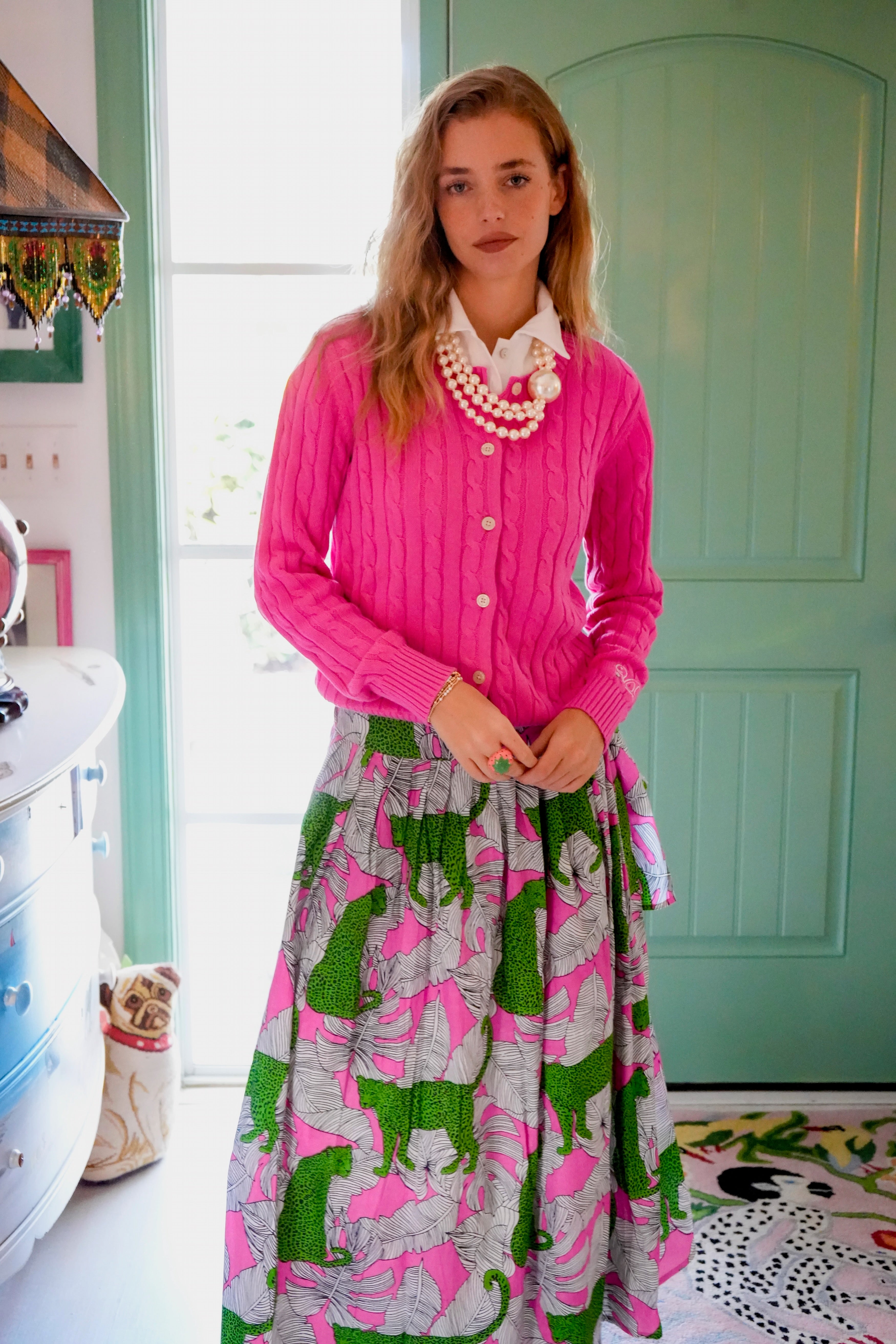 Woman wearing a pink cardigan and patterned skirt and jungle jag pink and green wrap skirt in a room with a green door.
