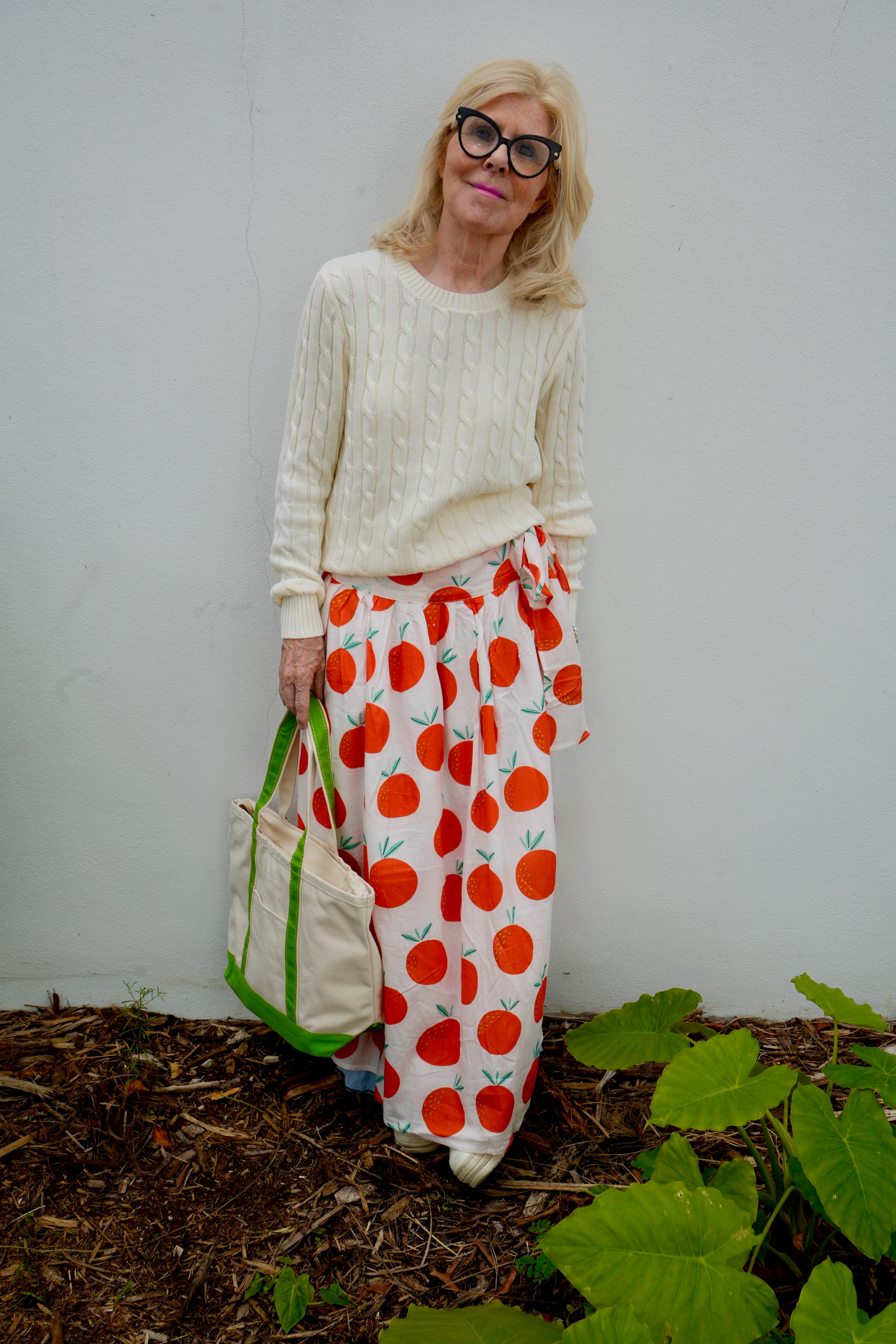Woman wearing a white sweater and orange patterned skirt standing in front of a white wall with greenery.